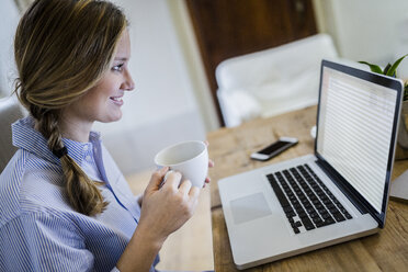 Smiling woman sitting at desk with cup of coffee and laptop - GIOF03646