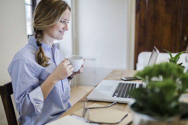Smiling woman sitting at desk with cup of coffee and laptop - GIOF03647