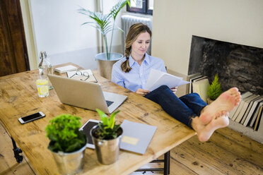 Frau sitzt am Schreibtisch zu Hause und legt die Füße hoch, um ein Dokument zu lesen - GIOF03652
