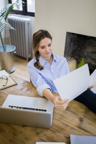 Frau sitzt am Schreibtisch zu Hause und legt die Füße hoch, um ein Dokument zu lesen, lizenzfreies Stockfoto