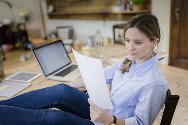 Frau sitzt am Schreibtisch zu Hause und legt die Füße hoch, um ein Dokument zu lesen - GIOF03656