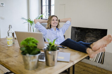 Lächelnde Frau sitzt am Schreibtisch zu Hause und legt die Füße hoch - GIOF03659