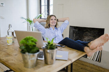 Smiling woman sitting at desk at home with feet up - GIOF03659