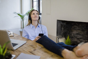 Smiling woman sitting at desk at home with feet up listening to music - GIOF03660