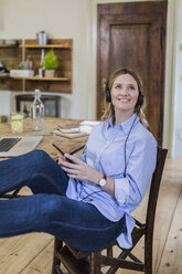 Smiling woman sitting at desk at home with feet up listening to music - GIOF03661