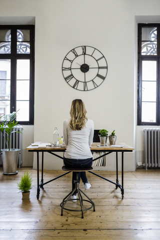 Rückansicht einer Frau, die zu Hause unter einer großen Wanduhr am Schreibtisch sitzt, lizenzfreies Stockfoto