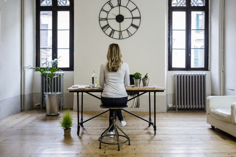 Rückansicht einer Frau, die zu Hause unter einer großen Wanduhr am Schreibtisch sitzt, lizenzfreies Stockfoto