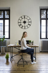 Portrait of smiling woman sitting at desk at home under large wall clock - GIOF03664