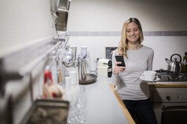 Smiling woman checking cell phone in kitchen - GIOF03678