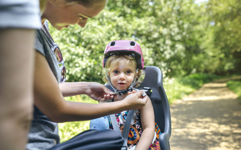 Little girl sitting on child seat for bicycle with her mother adjusting her helmet stock photo