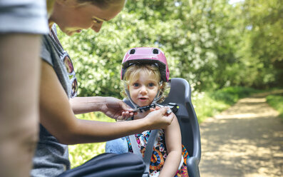 Kleines Mädchen sitzt auf einem Kindersitz für ein Fahrrad, während ihre Mutter ihren Helm einstellt - DAPF00821