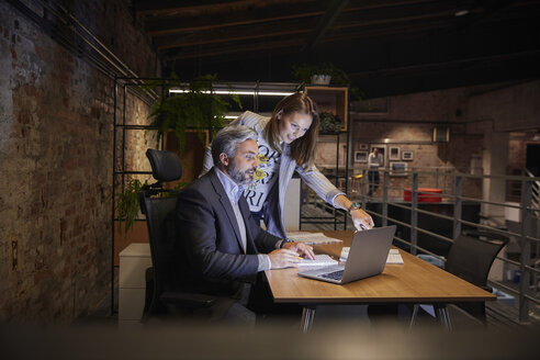 Businessman and woman sitting in modern office, looking at laptop - WESTF23796