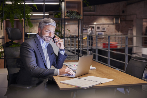 Mature businessman working in modern office, using laptop while talking on the phone stock photo