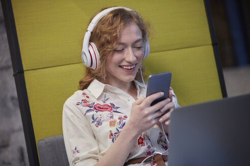 Junge Frau arbeitet in einem modernen, kreativen Büro, trägt Kopfhörer und benutzt ein Smartphone - WESTF23832