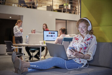 Junge Frau arbeitet in einem modernen, kreativen Büro, mit Laptop und Kopfhörern - WESTF23855