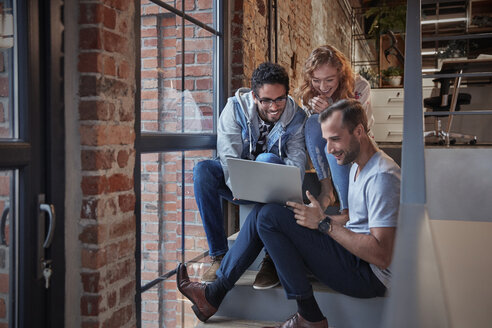 Young business people sitting on stairs in loft office using laptop - WESTF23859