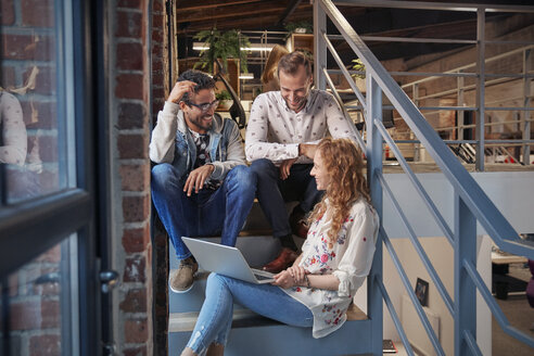 Young business people sitting on stairs in loft office using laptop - WESTF23860