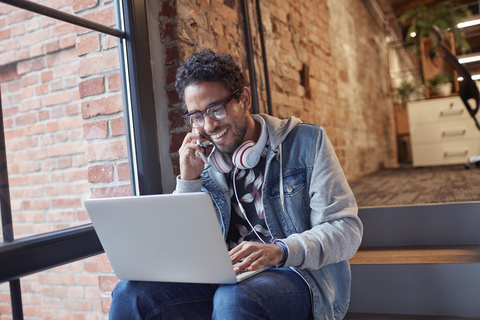 Junger Mann mit Kopfhörern und Laptop sitzt auf einer Bürotreppe und telefoniert, lizenzfreies Stockfoto