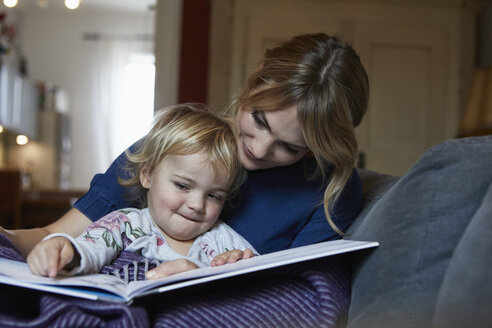 Mutter und kleine Tochter sitzen zu Hause auf der Couch und schauen ein Buch an - RBF06173