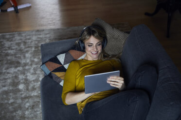 Porträt einer lächelnden Frau, die mit Kopfhörern und Tablet auf der Couch liegt - RBF06177