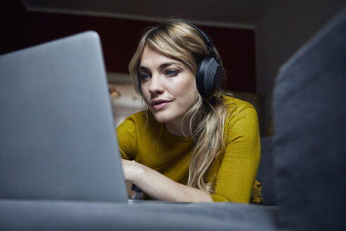 Porträt einer Frau, die mit Kopfhörern und Laptop auf der Couch liegt - RBF06178