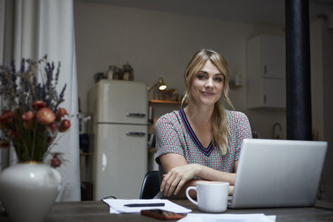 Portrait of smiling woman with laptop sitting at table in the kitchen - RBF06191