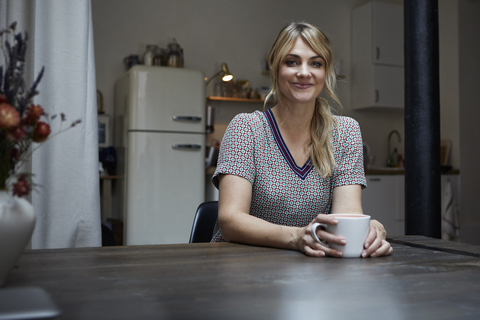 Portrait of smiling woman with cup of coffee sitting at table in the kitchen stock photo