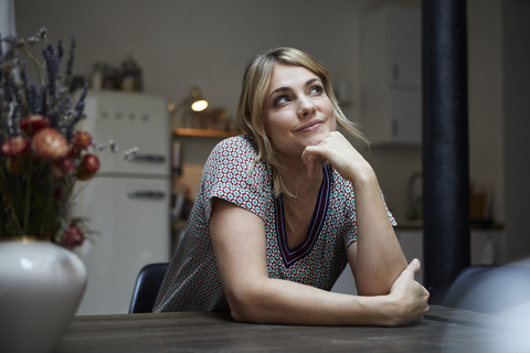 Portrait of smiling woman sitting at table in the kitchen thinking stock photo