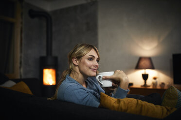 Porträt einer glücklichen Frau mit einer Tasse Tee, die sich abends zu Hause auf der Couch entspannt - RBF06202