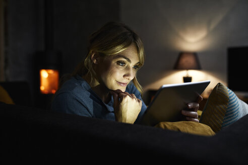Porträt einer lächelnden Frau, die abends auf der Couch zu Hause ein Tablet benutzt - RBF06205