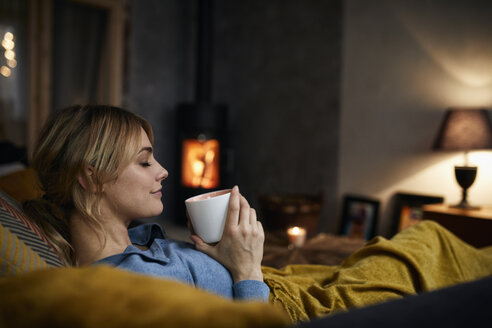 Lächelnde Frau mit einer Tasse Kaffee, die sich abends zu Hause auf der Couch entspannt - RBF06212