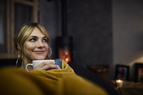 Porträt einer lächelnden Frau mit einer Tasse Kaffee, die sich abends zu Hause auf der Couch entspannt - RBF06218