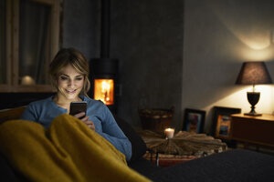 Porträt einer lächelnden Frau mit Smartphone, die sich abends auf der Couch entspannt - RBF06222