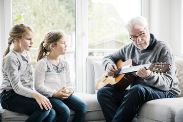 Two girls sitting on sofa listening to grandfather playing guitar - MOEF00519