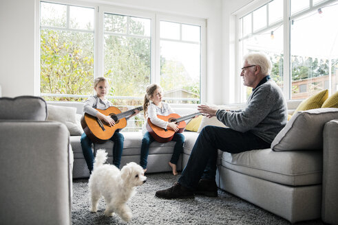 Zwei Mädchen auf dem Sofa spielen Gitarre mit Großvater und Hund - MOEF00522