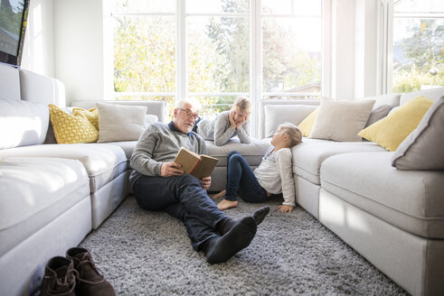 Two girls and grandfather reading book in living room - MOEF00526