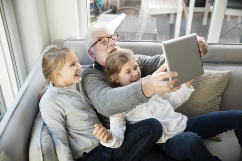 Zwei glückliche Mädchen und Großvater auf dem Sofa machen ein Selfie mit Tablet, lizenzfreies Stockfoto