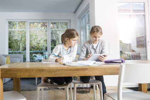 Zwei Mädchen machen gemeinsam Hausaufgaben am Tisch und benutzen ein Tablet, lizenzfreies Stockfoto