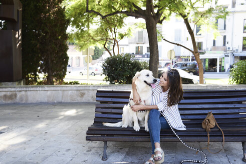Glückliche junge Frau sitzt mit ihrem Hund auf einer Bank in der Stadt - IGGF00319