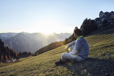 Österreich, Tirol, Rofangebirge, Wanderer sitzt bei Sonnenuntergang auf einer Wiese - RBF06224