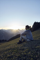 Österreich, Tirol, Rofangebirge, Wanderer sitzt bei Sonnenuntergang auf einer Wiese - RBF06225