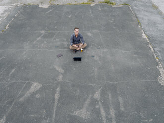 Young businessman with coffee to go and several mobile devices sitting on tarmac - KNSF03243