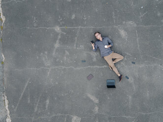 Young businessman with coffee to go lying on tarmac using mini tablet, quadcopter view - KNSF03244