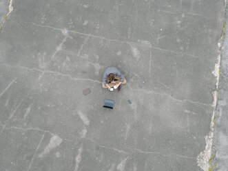 Young businessman with coffee to go and several mobile devices sitting on tarmac using tablet, quadcopter view - KNSF03246