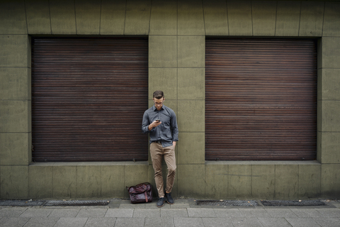 Young man leaning against house front using cell phone stock photo