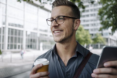 Porträt eines lachenden jungen Geschäftsmannes mit Coffee to go und Smartphone, lizenzfreies Stockfoto