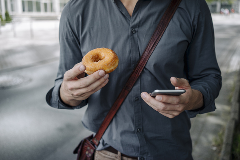 Hände eines Geschäftsmannes, der einen Donut und ein Smartphone hält, Teilansicht, lizenzfreies Stockfoto