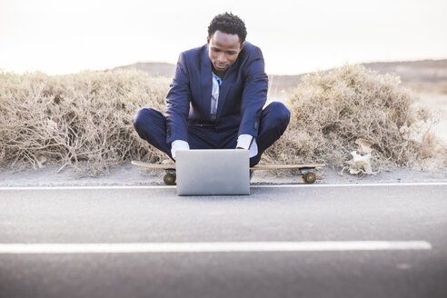 Spanien, Teneriffa, junger Geschäftsmann auf Skateboard sitzend und mit Laptop - SIPF01896