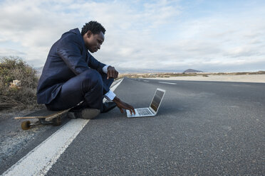 Spain, Tenerife, young businessman sitting on skateboard and using laptop - SIPF01907