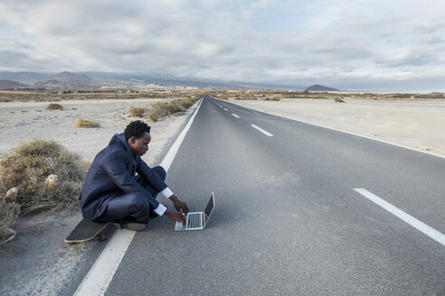 Spanien, Teneriffa, junger Geschäftsmann auf Skateboard sitzend und mit Laptop - SIPF01908